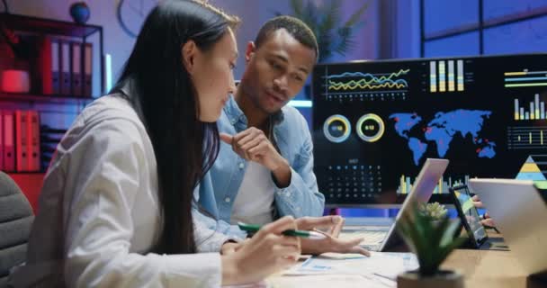 Atraktivní sebevědomí zkušený mladý asijský žena a africký Američan muž diskutovat o výsledcích finanční zprávy, zatímco pracuje společně v kanceláři v noci, front view — Stock video