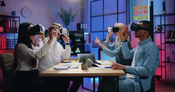 Красивая, уверенная в себе современная опытная команда разносторонних сотрудников, сидящих за столом в вечернем офисе и работающих в гарнитурах виртуальной реальности — стоковое видео