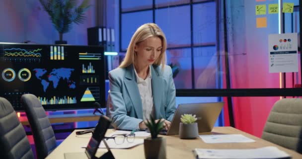 Krásná soustředěná cílevědomé kvalifikované blond obchodnice pracující na notebooku a dělat důležité poznámky v papírové zprávě ve večerní kanceláři, zblízka — Stock video