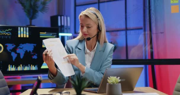 Прекрасная уверенная в себе опытная блондинка-бизнесмен в беспроводных наушниках сидит перед ноутбуком во время видеовстречи и объясняет отчет с графиками из вечернего офиса — стоковое видео