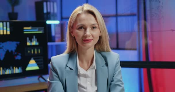 Jolie souriante satisfaite confiante qualifiée femme d'affaires blonde posant à la caméra dans un bureau moderne tout en faisant des heures supplémentaires le soir, vue de face — Video