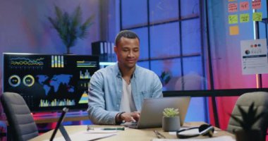 Yakışıklı, kendinden emin, deneyimli Afrikalı Amerikalı işçi akşamları modern ofiste bilgisayarda yazı yazıyor.