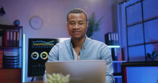 Närbild av snygg leende säker hårt arbetande skicklig afrikansk amerikansk man som sitter framför laptop på kvällen kontor och skriva på det med nöjda ansiktsuttryck — Stockvideo