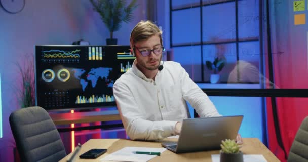 Koncept vzdálené práce, kde atraktivní sebevědomý motivovaný kvalifikovaný vousatý muž ve sluchátkách má videohovor s kolegou na počítači z večerní kanceláře — Stock video