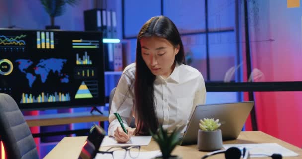 Likable concentrato fiducioso donna asiatica qualificato seduto sul posto di lavoro in ufficio sera e di lavoro con rapporti cartacei e informazioni sul computer portatile, vista frontale — Video Stock