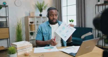 Yakışıklı, kendine güvenen, sakallı, kulaklıklı bir Afrikalı adam evde kameranın önünde oturmuş online konferans sırasında diyagramları anlatıyor.