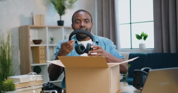Κοντινό πορτρέτο του όμορφου χαρούμενου μουσάτου Αφροαμερικανού που βγαίνει από το δέμα με τα νέα ακουστικά του και τα βάζει στο κεφάλι του. — Αρχείο Βίντεο