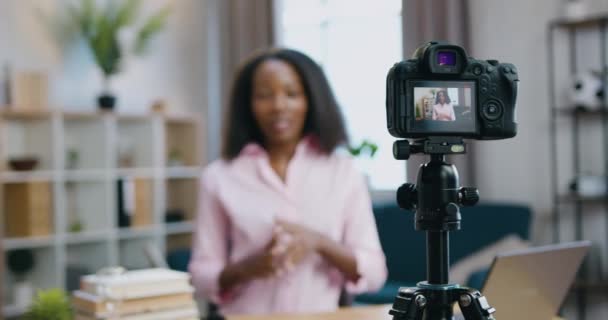 Fokusera på kamera där härlig positiv säker vänlig ung afrikansk amerikansk kvinna inspelning video för hennes internet anhängare från mysiga hem studio — Stockvideo