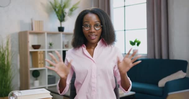 Вебкамера дивиться на чарівну позитивну впевненість молода афроамериканка в окулярах, які сидять за столом у домашній студії і розмовляють з аудиторією. — стокове відео