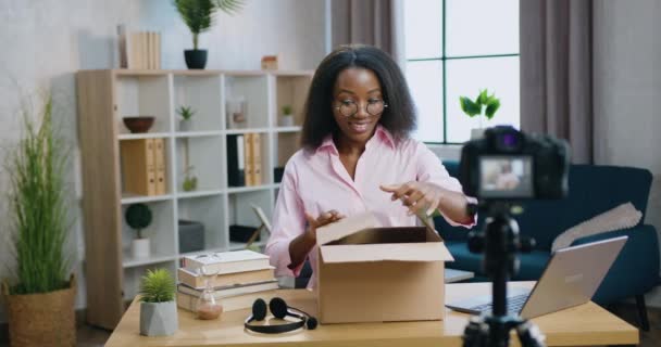 Unboxing proces waar charmante vrolijke glimlachende jonge Afrikaanse vrouw openen kartonnen doos met koptelefoon en zich verheugen tijdens het opnemen van video op camera thuis voor internet publiek — Stockvideo