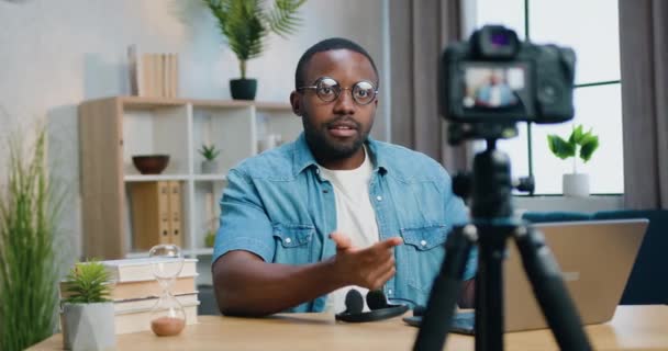 Atractivo blogger afroamericano amigable y confiado en gafas que graban video en cámara para su audiencia en Internet desde la acogedora oficina en casa, vista frontal — Vídeos de Stock