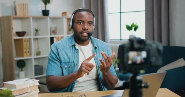 Gut aussehender selbstbewusster junger bärtiger Afroamerikaner im Headset, der ein neues Video für sein Internet-Publikum mit einer Videokamera auf dem heimischen Innenhintergrund aufnimmt — Stockvideo
