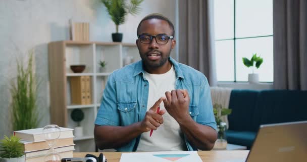 Beau positif confiant qualifié barbu afro-américain dans des lunettes assis devant la caméra à la maison et tenant une conversation vidéo avec le client — Video