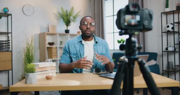 Atractivo amigable exitoso blogger africano barbudo en gafas filmando nuevo video para su audiencia con videocamera profesional en el fondo interior del hogar — Vídeos de Stock