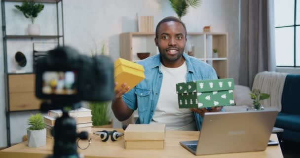 Vue de face sur beau sourire satisfait jeune homme afro-américain barbu qui tient des boîtes en carton avec des cadeaux dans les mains pendant l'enregistrement vlog sur la caméra pour la chaîne Internet — Video