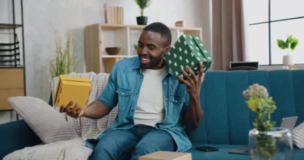 Yakışıklı, mutlu, mutlu, heyecanlı, sakallı, Afro-Amerikan adam. Evdeki kanepede oturuyor ve elinde iki hediye kutusu arasında seçim yapıyor. — Stok video