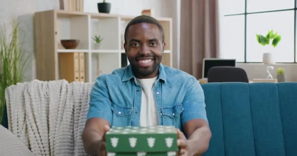 Attrayant sourire heureux insouciant jeune afro-américain barbu assis devant la caméra sur le fond intérieur de la maison et montrant belle boîte cadeau, vue de face — Video