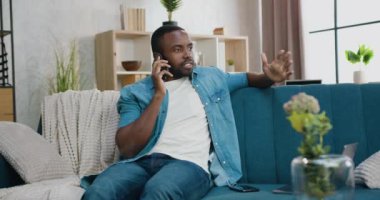 Çekici, kendine güvenen, genç, sakallı Afrikalı Amerikalı evdeki kanepede oturuyor ve telefon görüşmesi yapıyor.