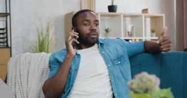 Çekici tatmin olmuş, gamsız, genç, sakallı Afrikalı adam modern oturma odasındaki kanepede oturuyor ve akıllı telefondan iyi bir ruh hali içinde konuşuyor.