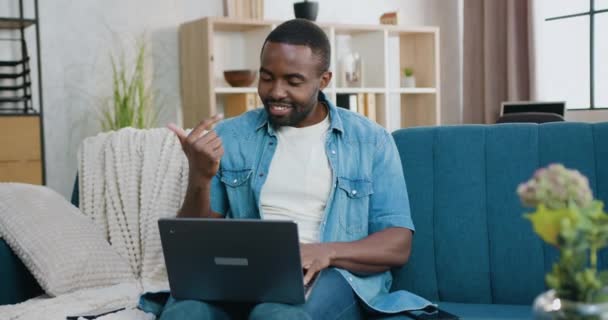 Портрет симпатичный улыбающийся счастливый расслабленный бородатый африканский американец, который сидит на диване дома во время видеочата с другом на компьютере, замедленная съемка — стоковое видео