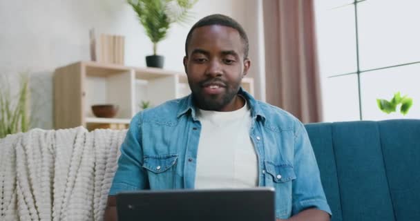 Otthon szabadidő koncepció, ahol jóképű mosolygós elégedett gondtalan fiatal szakállas afrikai srác ül a puha kanapén otthon alatt kellemes videó beszélgetés a számítógépen — Stock videók
