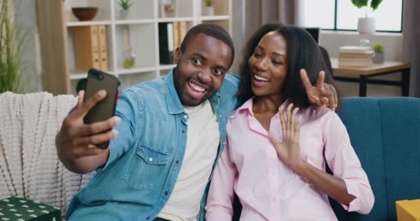 Attrayant joyeux heureux insouciant jeune couple afro-américain assis sur un canapé doux à la maison et faire selfie au téléphone avec des visages drôles, concept de loisirs en famille — Video