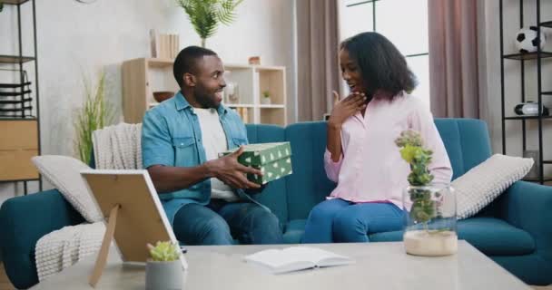 Bonita y sonriente enigmática pareja afroamericana joven sentados juntos en el sofá en casa y dándose cajas de regalo, disparando aceleradamente — Vídeos de Stock
