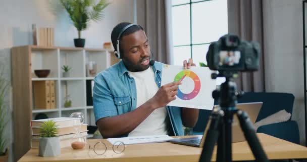 Ελκυστικό κίνητρο με αυτοπεποίθηση γενειοφόρος Αφροαμερικάνος άνδρας σε ακουστικά που καταγράφει βίντεο σε επαγγελματική κάμερα στο σπίτι και εξηγεί πολύχρωμο διάγραμμα για το κοινό του — Αρχείο Βίντεο