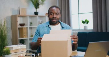 Kutuları açarken kameranın önünde oturan ve şaşırmış bir şekilde bakan sevimli, mutlu, mutlu, sakallı Afrikalı Amerikalı 'ya yakın durun.