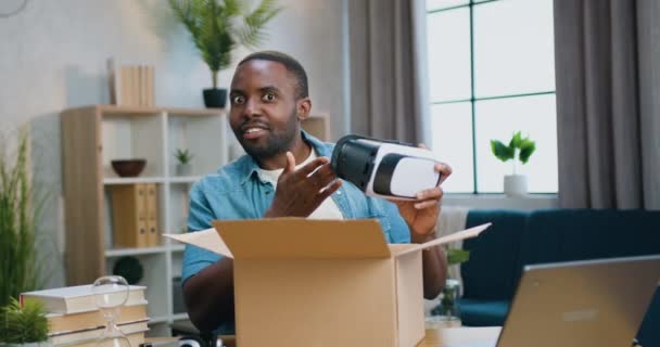 Atraente feliz sorrindo emocional barbudo americano africano tirando da caixa de papelão vr fone de ouvido e contando sobre suas oportunidades em câmera, acelerando a filmagem da câmera — Vídeo de Stock