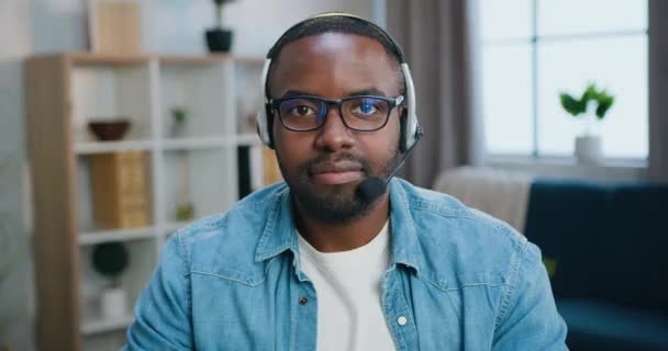 Porträtt av vacker leende nöjd ung skäggig afrikansk amerikan i glasögon i hörlurar som tittar in i kameran med vänliga ansiktsuttryck — Stockvideo