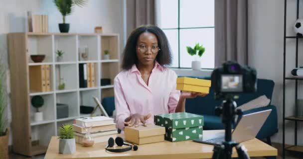 Blogging concept όπου γοητευτική χαμογελαστή χαρούμενη νεαρή Αφροαμερικανή που δείχνει κουτιά δώρων στην κάμερα κατά τη διάρκεια της εγγραφής του vlog για το διαδικτυακό της κανάλι — Αρχείο Βίντεο