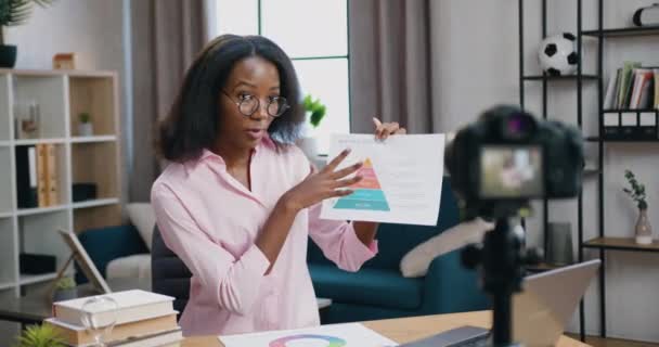 Χαριτωμένο θετική αυτοπεποίθηση ειδικευμένη Αφροαμερικανή γυναίκα κάθεται μπροστά από την κάμερα κατά τη διάρκεια της συνομιλίας βίντεο με τον πελάτη και εξηγώντας έκθεση με διάγραμμα, μακρινή έννοια εργασίας — Αρχείο Βίντεο