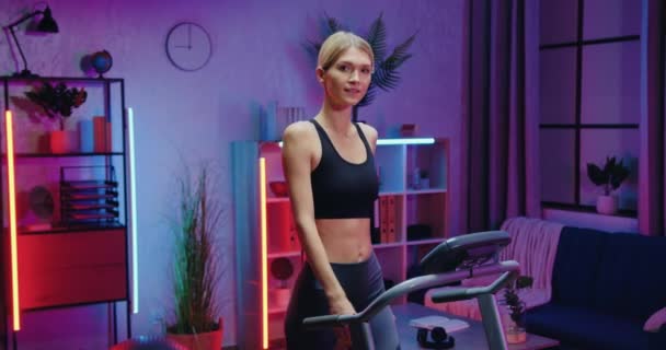Okouzlující pozitivní štíhlé sportovní mladá žena ve sportovním oblečení pózovat na kameře při šlapání na běžecký pás během večerního domácího cvičení, zpomalený pohyb — Stock video