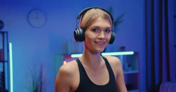 Крупним планом портрет усміхненої активної стрункої молодої жінки в навушниках, яка дивиться в камеру під час тренувань на біговій доріжці під час вечірніх тренувань вдома — стокове відео