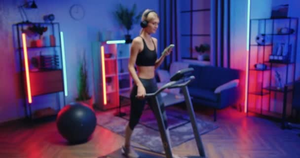Sporty lifestyle concept where attractive positive active slim young woman in sportswear in headphones walking on treadmill at home in the evening and looking into camera with sincerely smile — Stock Video