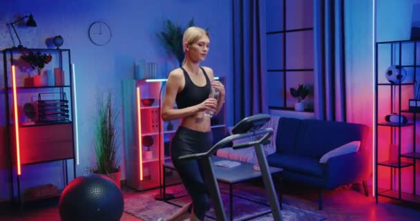 Goed uitziende actieve pasvorm slanke jonge vrouw in training kleding wandelen op loopband en drinkwater tijdens thuis avond workout, gezonde levensstijl concept — Stockvideo