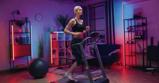 Вечірній домашній тренувальний концепт, де приваблива спортивна активна струнка блондинка в спортивному одязі робить кардіо вправи на біговій доріжці, повільний рух — стокове відео