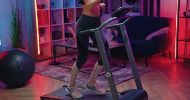 Привлекательная улыбающаяся активная спортивная молодая женщина в тренировочной одежде делает беговые упражнения на беговой дорожке во время домашней вечерней тренировки — стоковое видео