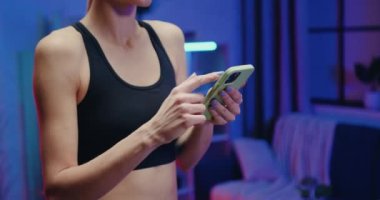 Siyah elbiseli, etkin ve zayıf bir kadın koşu bandına basıyor ve akşam antrenmanı sırasında cep telefonunu kullanıyor.