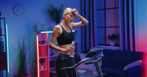 Gyönyörű sportos illik karcsú fáradt fiatal nő sportruházat ivóvíz üvegből, miközben a lépés gyakorlatok futópadon hangulatos esti szobában — Stock videók