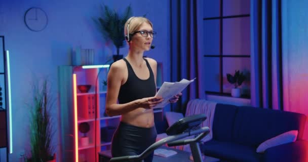 Lovely confident sporty slender young woman in headset with mic revisioning documents,talking with interlocutor during stepping on treadmill at home in the evening — Stock Video