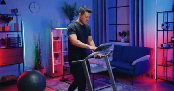 Привлекательный уверенный в себе спортивный азиатский мужчина в спортивной одежде устанавливает скорость на контрал-панели, делая шаговые упражнения на беговой дорожке дома вечером — стоковое видео