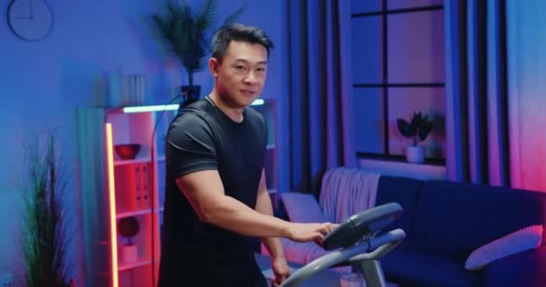 Красивый улыбающийся активный спортивный азиат в тренировочной одежде ходит по беговой дорожке и показывает большие пальцы на фоне ночной комнаты — стоковое видео