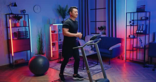 Активная концепция образа жизни, где привлекательный спортивный мускулистый молодой азиат в тренировочной одежде работает на беговой дорожке во время вечерних домашних тренировок — стоковое видео