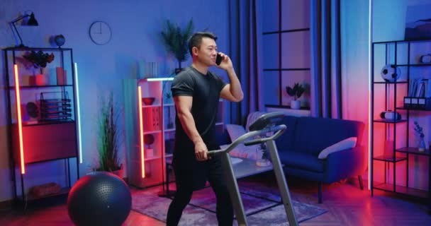 Snyggt leende nöjd aktiv muskulös asiatisk kille i sportkläder som tränar på löpband och pratar i mobilen under kvällsträning hemma — Stockvideo
