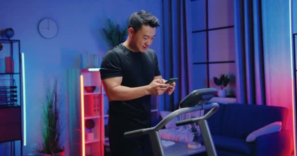 Привлекательный счастливый сверхрадостный активный спортивный азиатский мужчина в спортивной одежде, получающий хорошие новости на смартфоне во время занятий на беговой дорожке во время вечерней домашней тренировки — стоковое видео