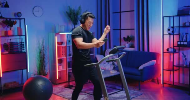 Sympathisch sportlich aktiver asiatischer Mann in Sportbekleidung, der auf dem Laufband läuft und während des abendlichen Heimtrainings über Kopfhörer Musik von der Handy-Playlist hört — Stockvideo