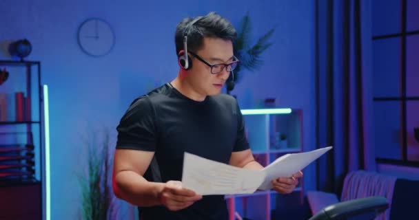 Attractive confident active sportive asian businessman in sportswear wears headphones with mic talking with interlocutor, reviewing reports during home workout in the evening — Stock Video