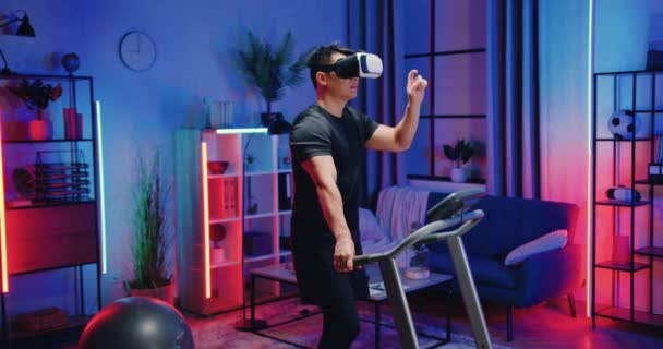 Atraente confiante ativo desportivo asiático cara no sportswear andando na esteira rolante e aplicando realidade virtual fone de ouvido durante o treino em casa à noite, tecnologia moderna e conceito de estilo de vida — Vídeo de Stock